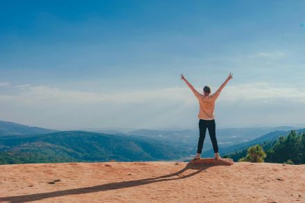 A man raises his arms in triumph on a rocky mountain summit overlooking a vast landscape