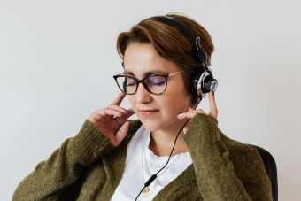Content glad female wearing eyeglasses and headphones listening to good music and touching headset while sitting with eyes closed against white wall