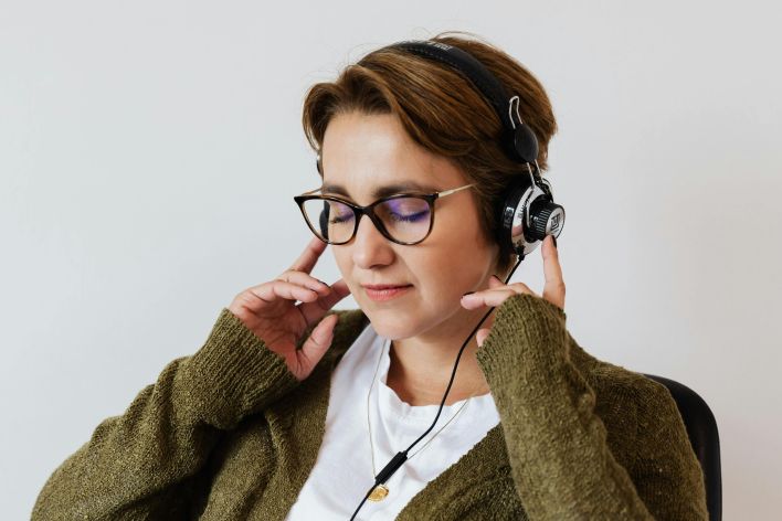 Content glad female wearing eyeglasses and headphones listening to good music and touching headset while sitting with eyes closed against white wall